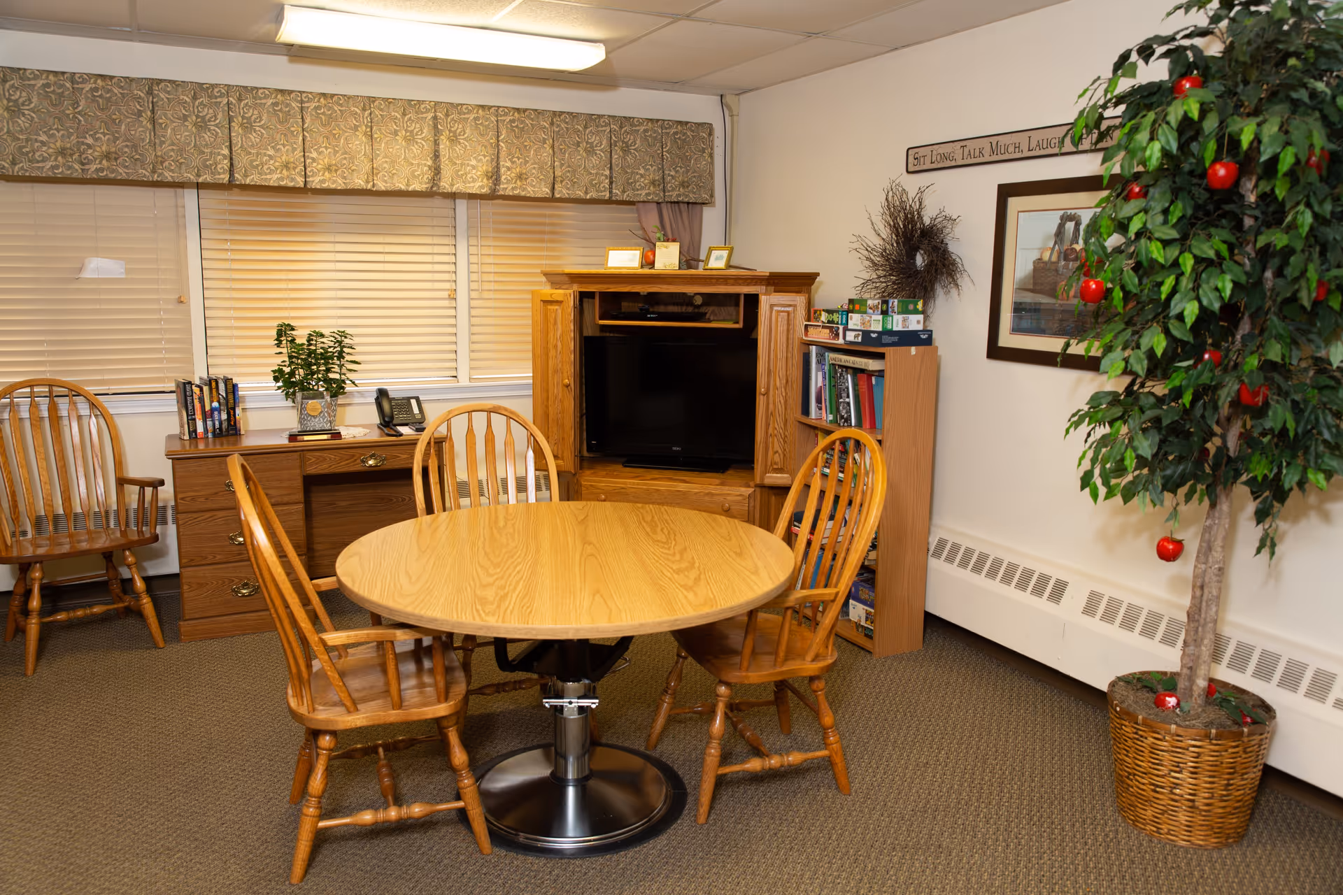 A cozy room with a round wooden table surrounded by four wooden chairs. In the background, there is a wooden desk with books, a plant, and a telephone on it. Next to the desk is a wooden entertainment center with a television. A bookshelf with board games and books is placed beside the entertainment center. A large potted artificial tree with red apples is on the right side of the room. The walls are decorated with a framed picture, a wreath, and a sign that reads 'Sit Long, Talk Much, Laugh Often.' The room has carpeted flooring and windows with closed blinds and a patterned valance.