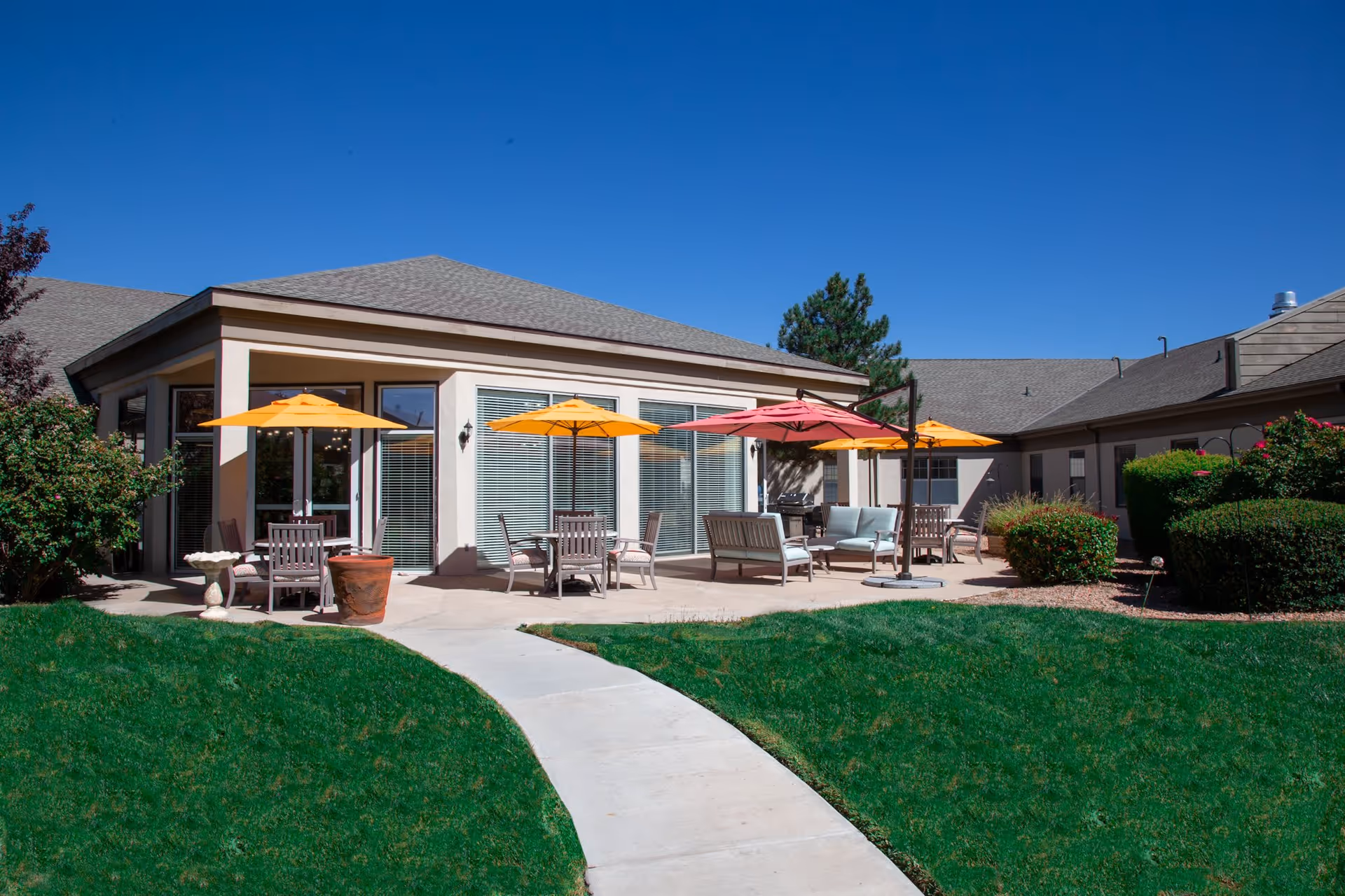 Outdoor patio area of a senior living facility with multiple seating arrangements including chairs and sofas under colorful umbrellas. The patio is surrounded by green grass, bushes, and a clear blue sky overhead.