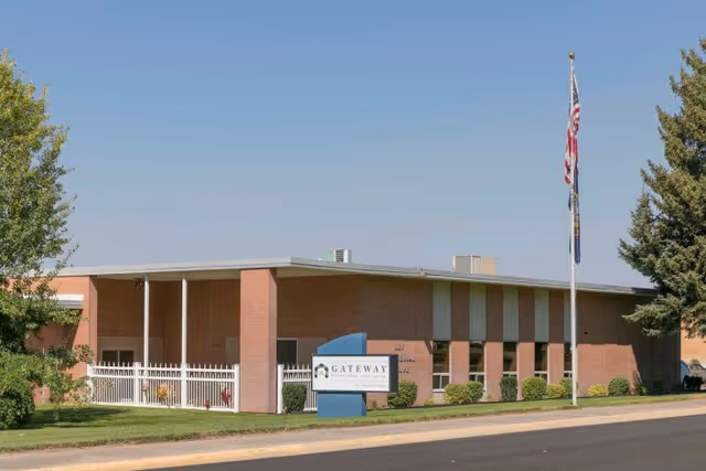 Exterior view of Gateway Transitional Care Center, a single-story brick building with a flat roof, surrounded by green grass and trees. There is a sign in front of the building displaying the facility's name, and two flagpoles with the American flag and another flag. The sky is clear and blue.