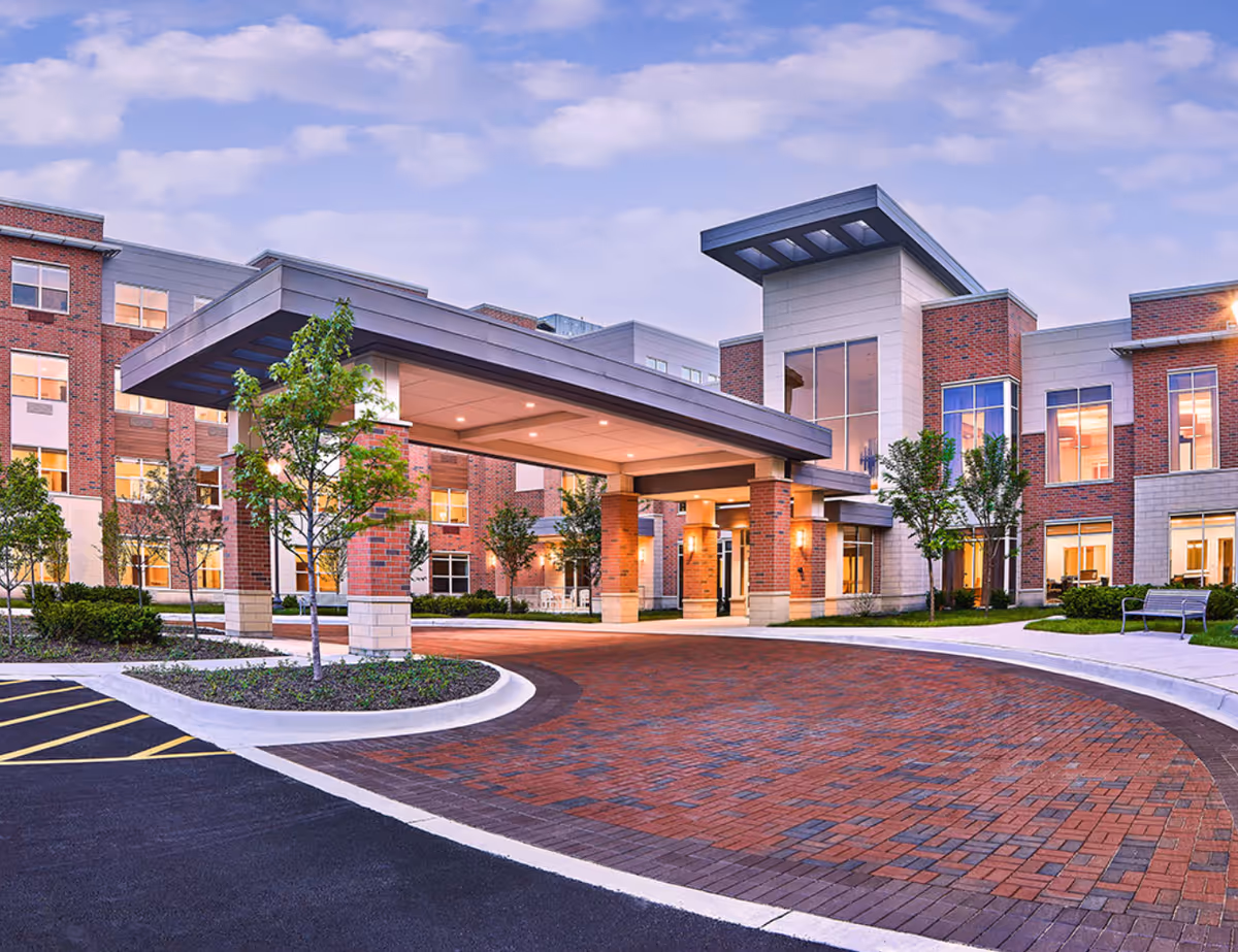 Exterior view of Oak Trace Health Center showing a modern building entrance with a covered drop-off area, brick and stone facade, large windows, landscaped greenery, and a paved driveway under a partly cloudy sky.