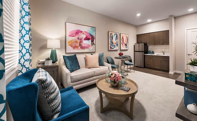 Bright furnished living room with teal-accented armchair and sofa, a round coffee table, wall art, and a small kitchenette in the background.