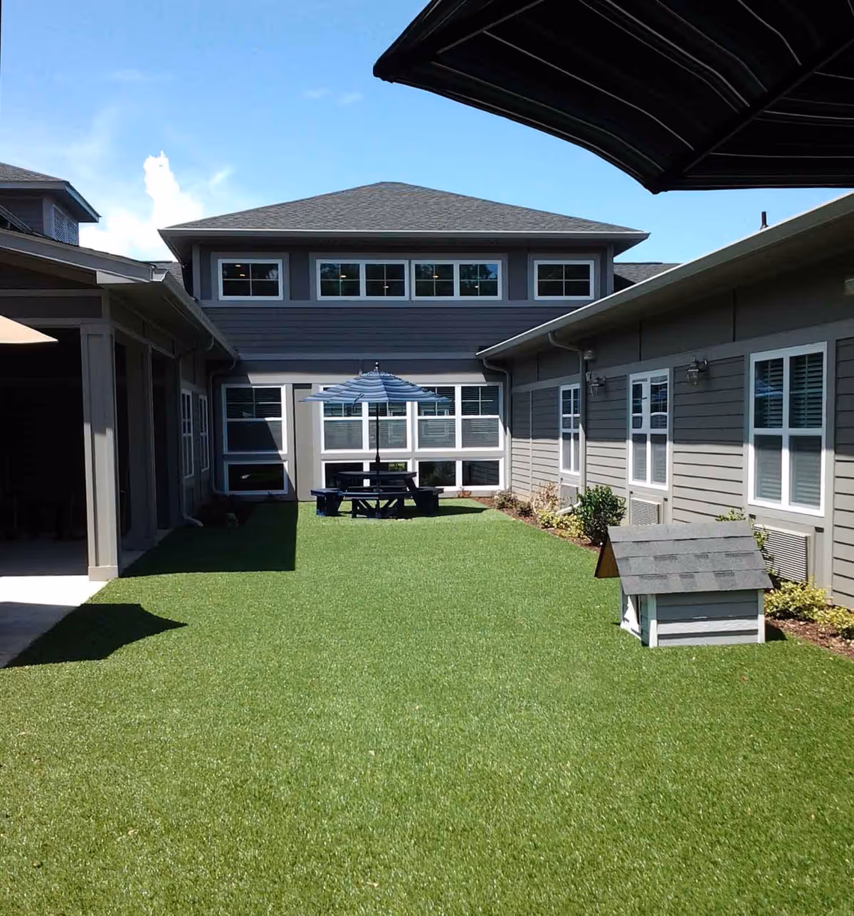 Outdoor courtyard area at Northlake House with artificial grass, a picnic table with a blue and white striped umbrella, and a small doghouse. The courtyard is surrounded by gray buildings with white-trimmed windows under a clear blue sky.