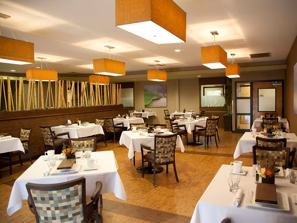 A well-lit dining room in Blue Point Healthcare Center with multiple tables covered in white tablecloths, each set with cups, glasses, and small flower arrangements. The room features modern square pendant lights hanging from the ceiling and patterned chairs around the tables. A wooden divider with vertical sticks separates part of the room, and there are framed artworks on the walls.