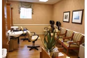 A waiting room with several striped cushioned chairs lined up against the wall, a small table with a potted plant in the foreground, and two hair dryer chairs near the back wall under a window. The room has wood flooring and beige wallpaper with framed pictures on the wall.