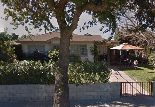 Front exterior view of a single-story residential building with a small garden and patio area. There is a large tree in the foreground, a low concrete block wall, and a metal gate. The patio has an umbrella and outdoor seating with some colorful decorations.