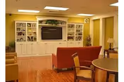 A cozy living room area in a nursing and rehabilitation center featuring a large built-in white entertainment center with a flat-screen TV, shelves with decorative items, and cabinets. The room has wooden flooring, a red sofa, a round table with chairs, and warm yellow walls with ceiling lights.