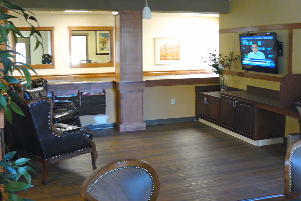 A cozy living room area with wooden flooring, dark leather armchairs, a wall-mounted flat-screen TV, wooden cabinets, and decorative plants. The walls are painted yellow with wooden trim, and there are framed pictures and mirrors on the walls.