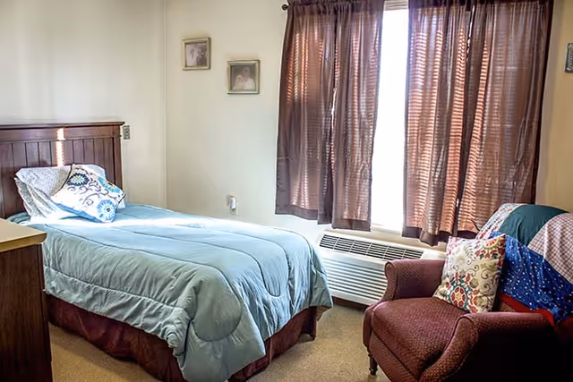 A small bedroom with a single bed covered in a light blue comforter and a decorative pillow. Next to the bed is a wooden nightstand. There is a maroon armchair with a colorful quilt and a patterned pillow near a window with brown curtains. Two small framed pictures hang on the wall above the bed.