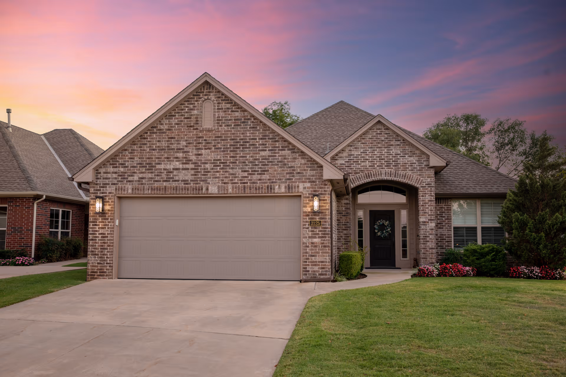 Front exterior view of a single-story brick house with a two-car garage, a black front door with a wreath, well-maintained lawn, and colorful flower beds under a vibrant sunset sky.