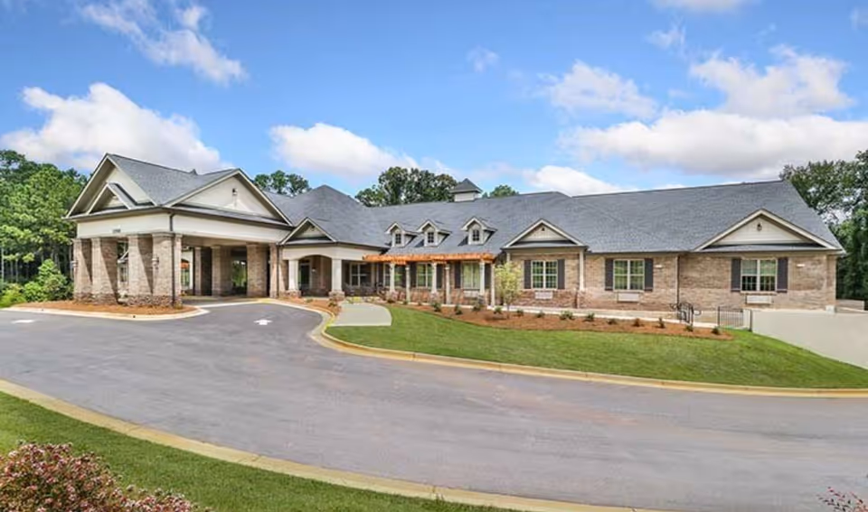 Exterior front view of a single-story brick senior living building with a porte-cochère, driveway and landscaped lawn under a blue sky.
