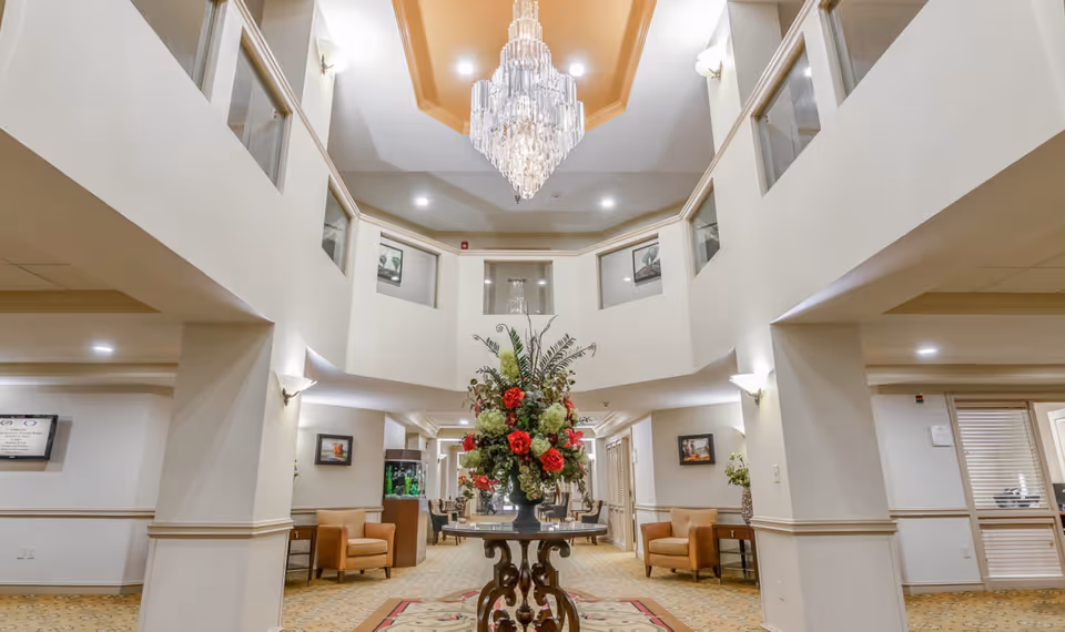 Two-story senior living lobby with a central floral arrangement on a table, chandelier overhead, and seating areas along the sides.