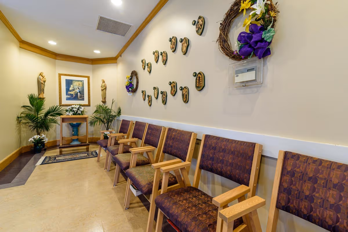A calm interior waiting area with a row of wooden chairs, religious statues and artwork, potted plants, and decorative wall wreaths.