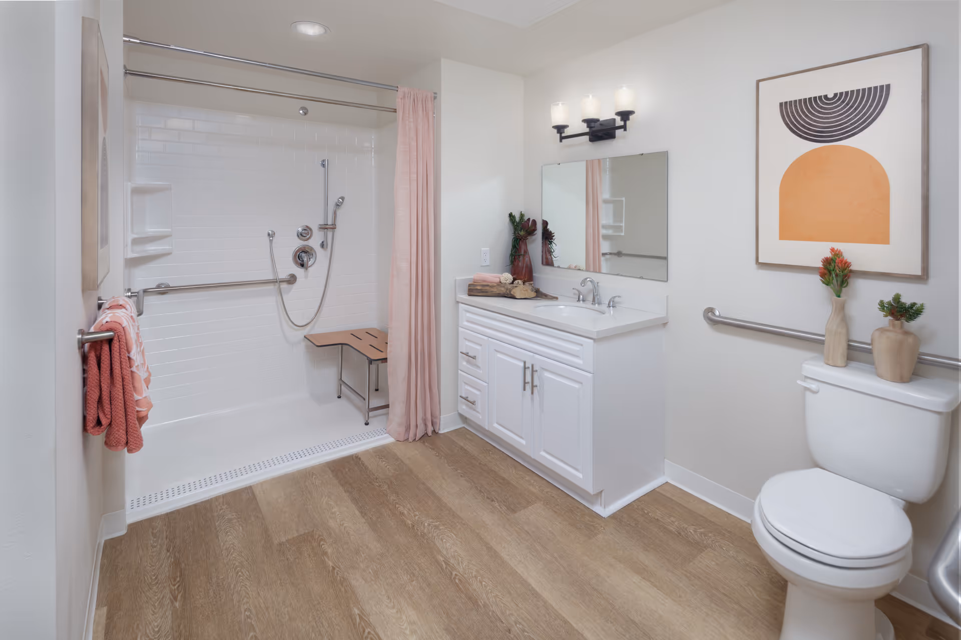 A clean and modern bathroom featuring a walk-in shower with a foldable wooden bench and a pink shower curtain. The bathroom has a white vanity with a sink, a large mirror above it, and a three-light wall fixture. There is a toilet with two small vases containing plants on top, a towel rack with pink towels, and a piece of abstract wall art with orange and black shapes. The floor is wood-style vinyl, and grab bars are installed near the shower and toilet for accessibility.