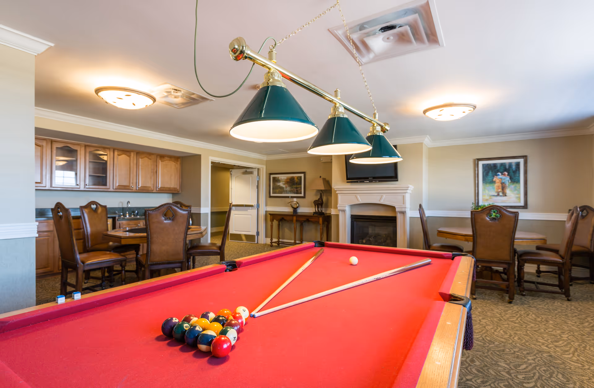A recreational room with a red pool table in the foreground, two pool cues resting on the table, and a rack of billiard balls set up. The room has a cozy ambiance with a fireplace, a TV mounted above it, wooden chairs and tables, and warm lighting fixtures on the ceiling. There are framed paintings on the walls and cabinetry with a sink in the background.