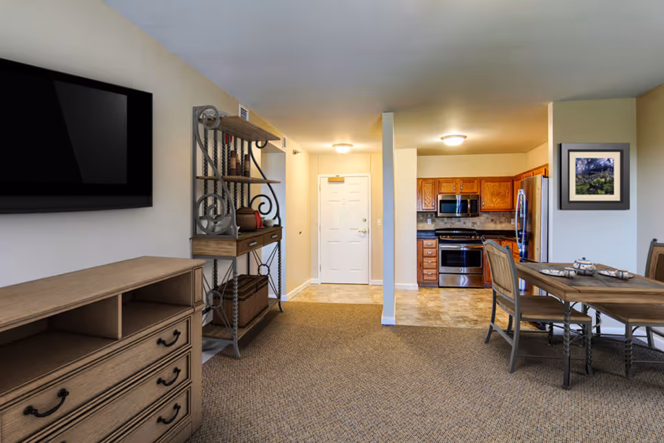 Interior view of a senior living community apartment showing a living area with a wall-mounted TV, a wooden dresser, a decorative metal and wood shelving unit, a dining table with chairs set for tea, and a kitchen with wooden cabinets, stainless steel appliances, and a tiled floor. The entrance door is visible in the background.