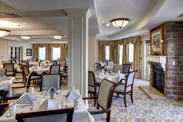 A spacious dining room with multiple tables set with white tablecloths, folded napkins, cups, glasses, and silverware. The room features upholstered chairs, large windows with beige curtains, and a stone fireplace with a framed painting above it. The carpet has a patterned design and the ceiling has several light fixtures.