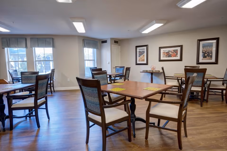 A bright dining room with wooden floors and multiple square wooden tables, each surrounded by four cushioned chairs with wooden frames. The room has large windows with light blue valances allowing natural light to enter. The walls are decorated with framed pictures, and the ceiling has rectangular fluorescent lights.