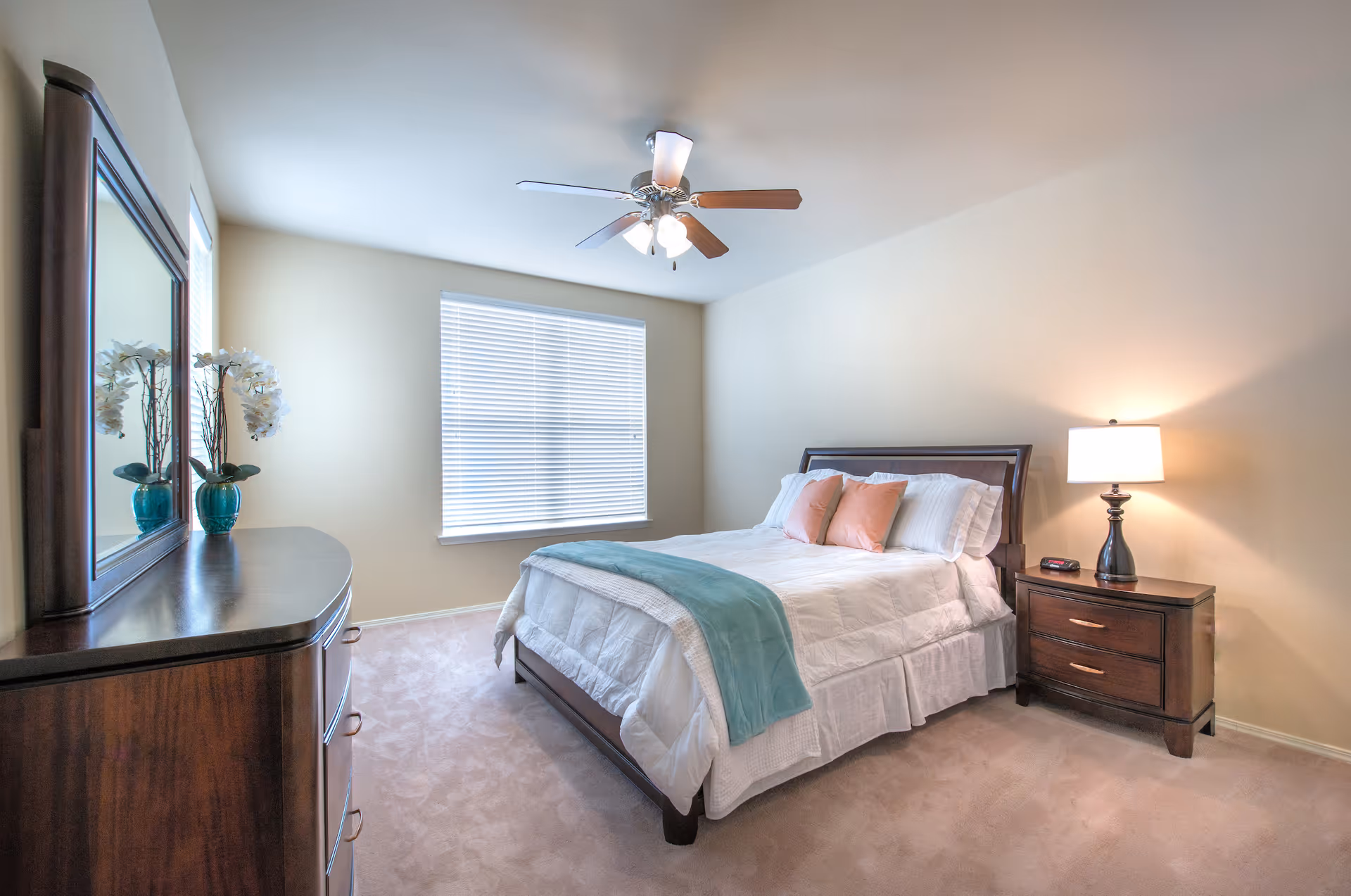 Bright, neatly staged bedroom with a bed, nightstand and lamp, dresser with mirror, and a ceiling fan.