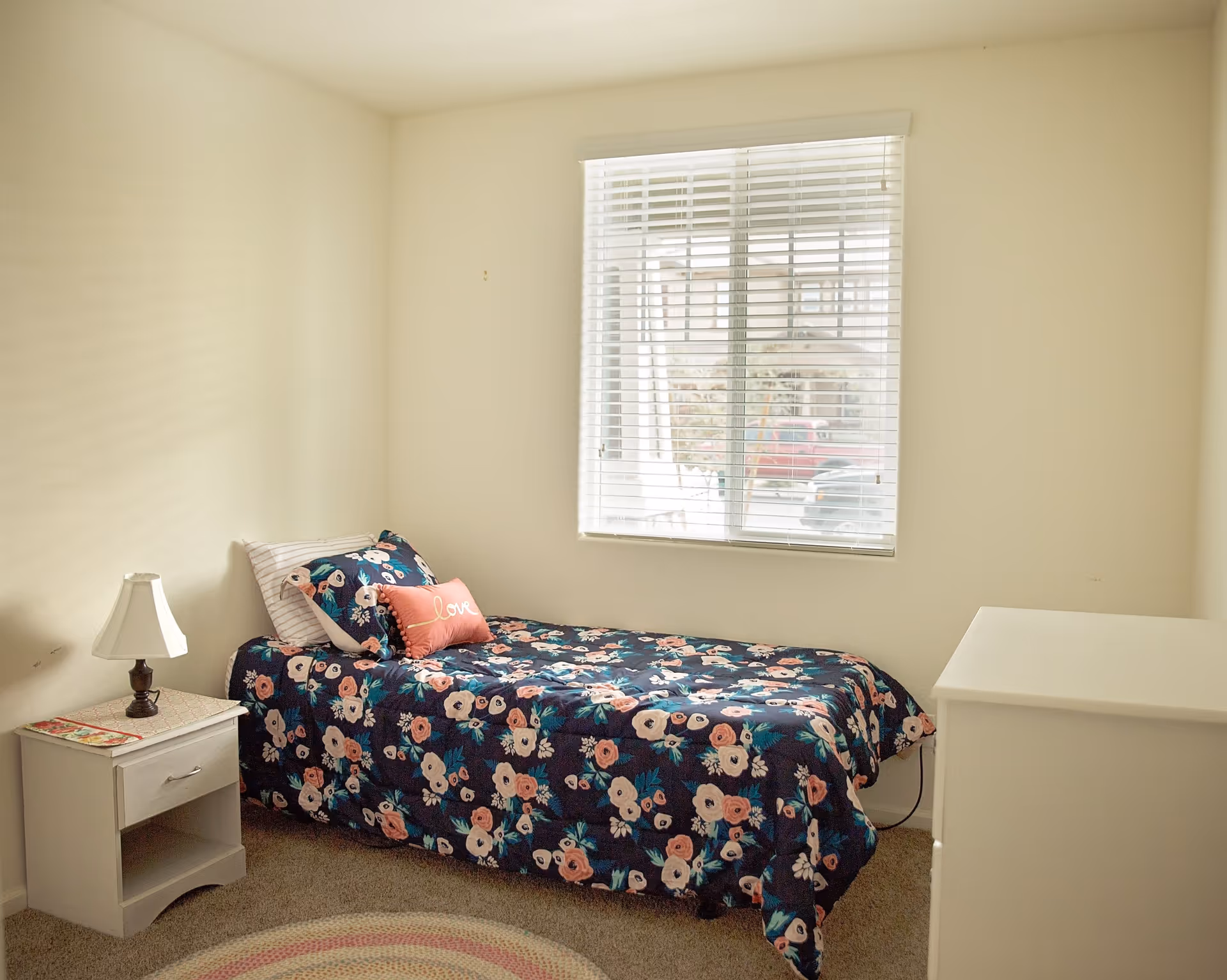 A small bedroom with a single bed covered in a dark floral bedspread and pillows, including one with the word 'love'. There is a white nightstand with a lamp on the left side of the bed and a white dresser on the right side. A window with white blinds lets in natural light, and the walls are painted a light beige color.