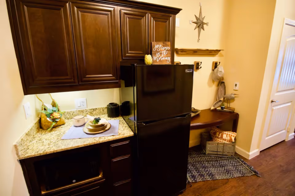 A compact kitchen area with dark wooden cabinets, a black refrigerator, and a granite countertop. On the countertop, there is a small plant, a cup, and a plate with a decorative item. Next to the refrigerator is a small wooden table with decorative items, a basket with pillows underneath, and a wall-mounted coat rack with a hat and a star-shaped wall decoration above.