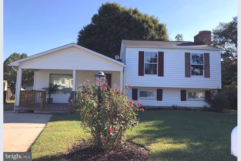 Exterior view of a white two-story house with brown shutters, a front porch with wooden railings, a driveway, and a garden with a flowering bush and a lamp post in the front yard under a clear blue sky.