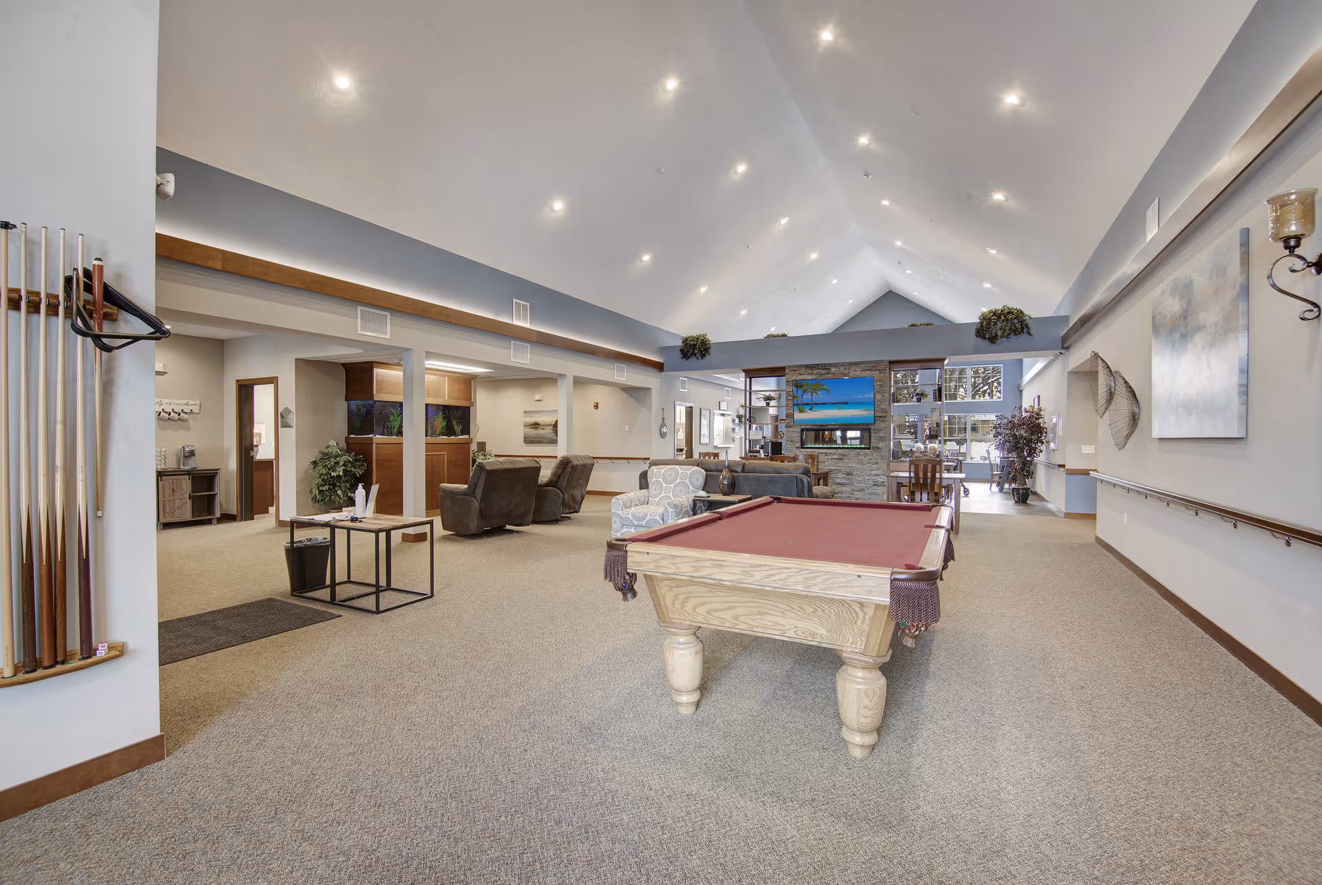 A spacious common area in a senior living facility featuring a pool table with a red felt top in the foreground. The room has high vaulted ceilings with recessed lighting, carpeted floors, and neutral-colored walls. There are several comfortable chairs and sofas arranged around a stone fireplace with a flat-screen TV mounted above it. The room also includes decorative plants, wall art, and a rack with pool cues on the left side.