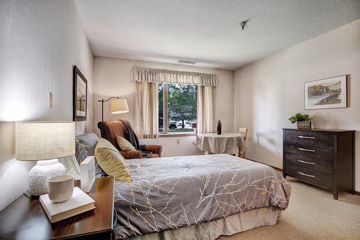 A cozy bedroom with a bed covered in a gray and white patterned comforter, a wooden nightstand with a lamp, cup, and book, a comfortable armchair with a floor lamp beside it, a small round table with two chairs near a window with beige curtains, and a dark wooden dresser with a plant and framed picture on the wall above it.
