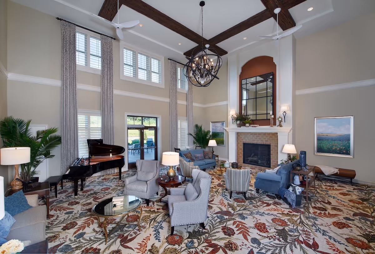 A spacious and elegant living room with high ceilings and exposed wooden beams. The room features a grand piano, multiple seating areas with armchairs and sofas, a large patterned rug, a fireplace with a large mirror above it, and tall windows with long curtains. There are also several lamps and framed paintings on the walls, along with some indoor plants.