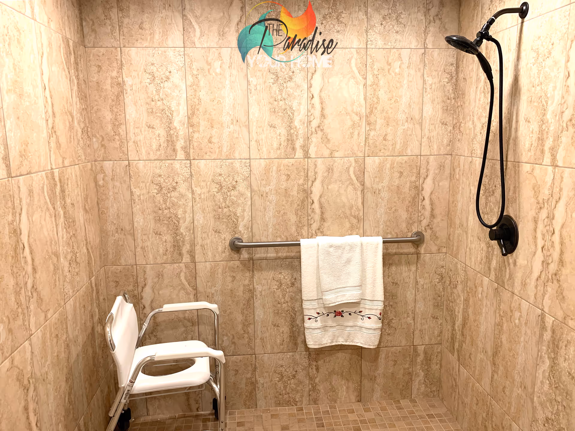 A tiled shower area with beige marble-like tiles on the walls and small square tiles on the floor. There is a white shower chair with armrests and wheels positioned in the corner. A metal grab bar is mounted on the wall with a white towel hanging on it. A black handheld showerhead is attached to the wall on the right side. The logo of The Paradise Assisted Living Home is visible at the top center of the image.
