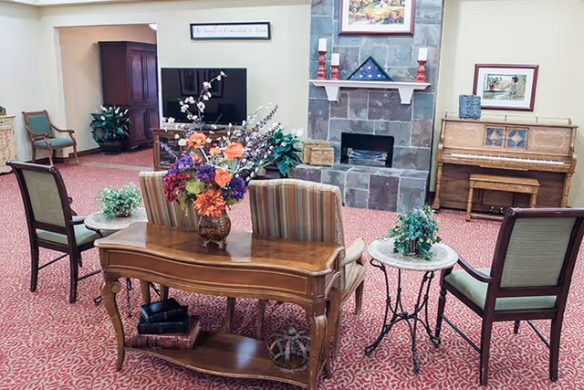 A cozy living room area with a red patterned carpet, a stone fireplace with candles and a folded American flag on the mantel, a wooden piano with a framed picture above it, a TV on a stand, and several chairs arranged around small tables. A wooden console table with a vase of colorful flowers is in the foreground.