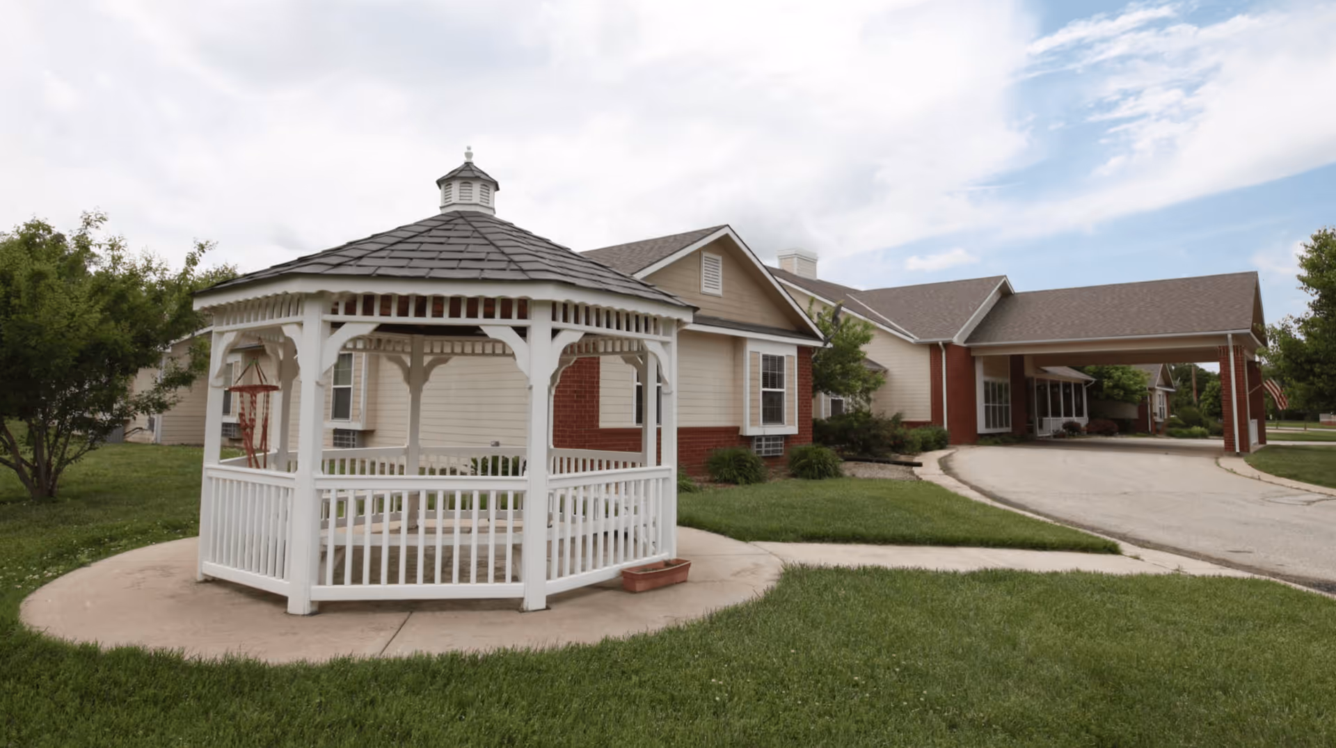 A white wooden gazebo on a lawn in front of a single-story assisted living building with a covered drive-through entrance.