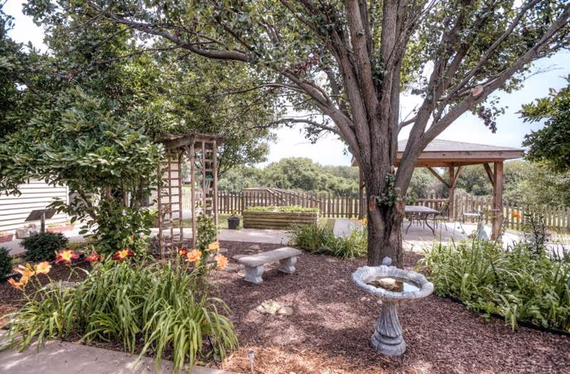 A peaceful outdoor garden area with a large tree, a birdbath, a stone bench, colorful flowers, and a wooden pergola with seating. There is a wooden fence in the background and lush greenery surrounding the space.