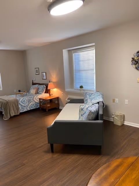 A cozy bedroom with a single bed covered in patterned blue and white bedding, a wooden nightstand with a lamp, and a gray sofa with matching cushions. The room has wooden flooring, a window with blinds, and neutral-colored walls.