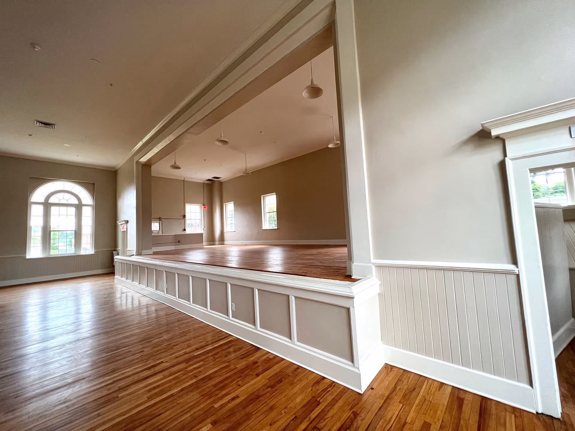 An empty room with polished wooden floors and beige walls featuring a raised stage area. The room has several windows allowing natural light to enter, and pendant lights hang from the ceiling. The space appears clean and well-maintained.