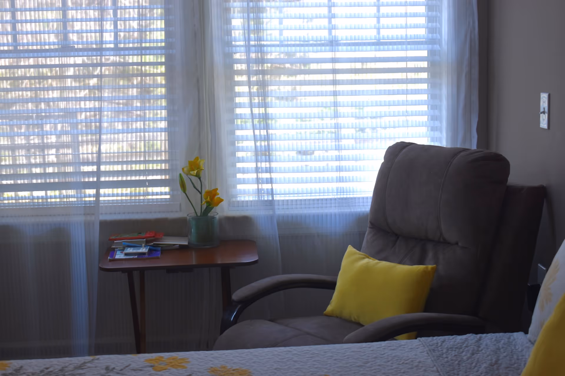 A cozy corner of a room with a large window covered by white blinds and sheer curtains. In front of the window is a small wooden side table with a green vase holding yellow flowers and some books or magazines. Next to the table is a comfortable gray armchair with a yellow pillow. Part of a bed with a white quilted bedspread featuring yellow floral patterns is visible in the foreground.