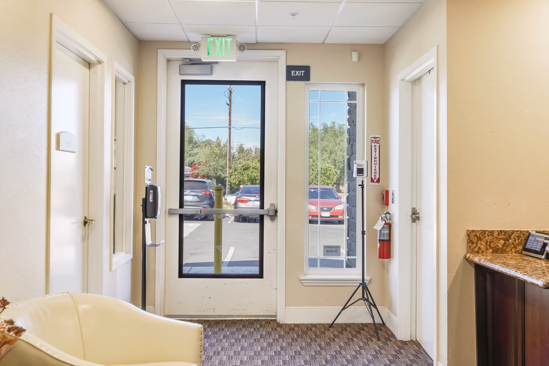 Interior view of an exit door area in a senior living facility with a glass door showing parked cars outside. There is a beige armchair in the foreground, a hand sanitizer dispenser on a stand, a fire extinguisher mounted on the wall, and a granite countertop on the right side.
