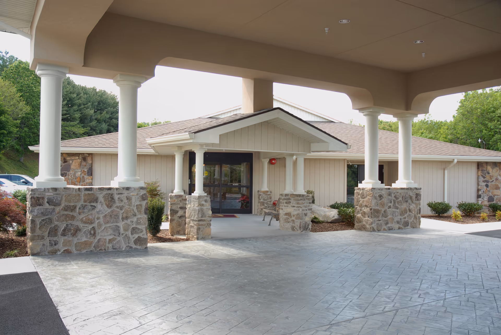 Covered entrance area of a single-story building with stone pillars and white columns, leading to glass double doors. The building is surrounded by greenery and has a paved driveway.