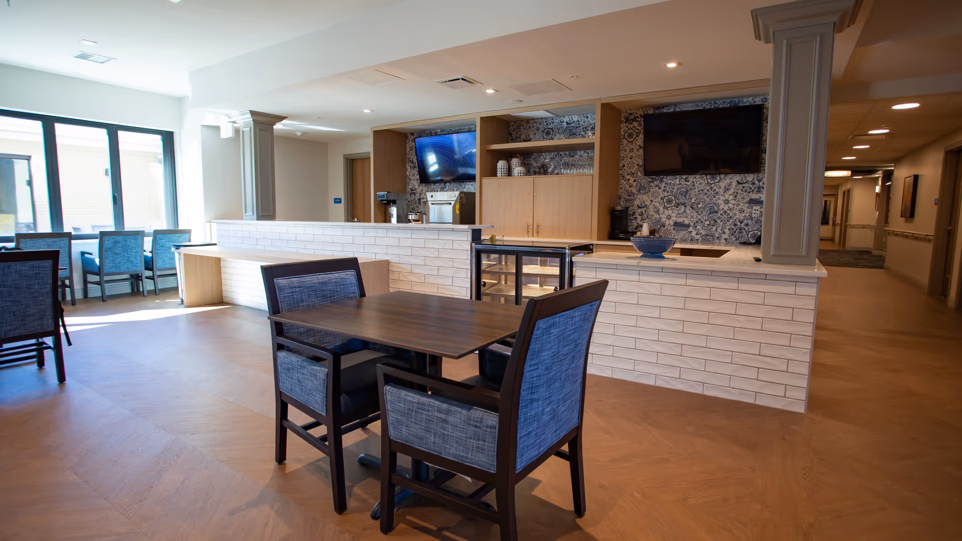 Bright communal dining area with wooden tables and upholstered chairs facing a service counter and TV screens.