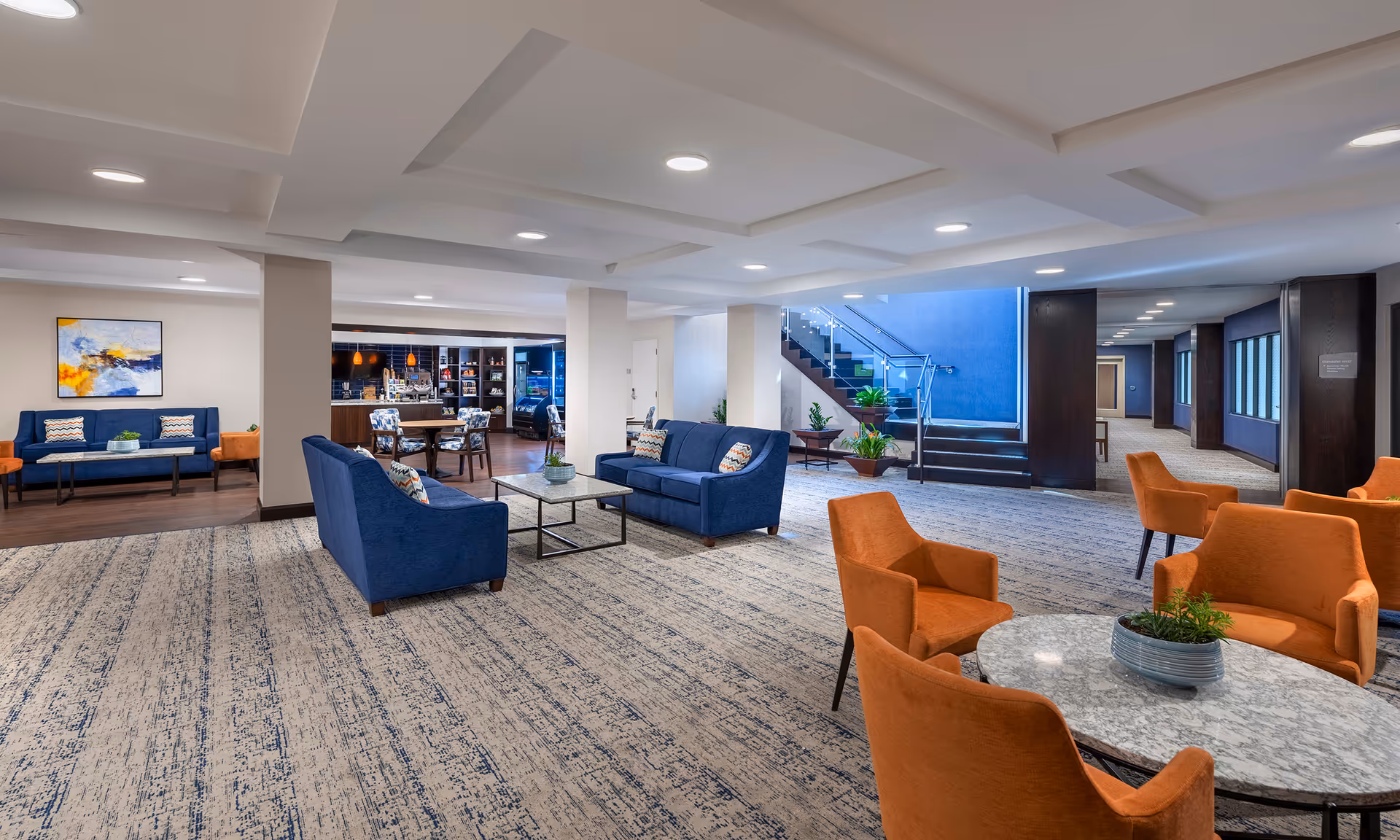 A spacious and modern common area with blue and orange upholstered chairs and sofas arranged around marble and glass tables. The room features a patterned carpet, recessed ceiling lights, a staircase with glass railing, and a small café or snack area in the background with shelves and hanging pendant lights.