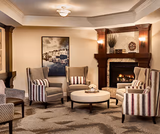 A cozy living room area with four upholstered armchairs arranged around a round coffee table. The chairs have striped cushions, and there is a lit fireplace with a wooden mantel in the background. Above the fireplace is a decorative piece and two wall sconces providing warm lighting. A framed picture hangs on the wall to the left of the fireplace.