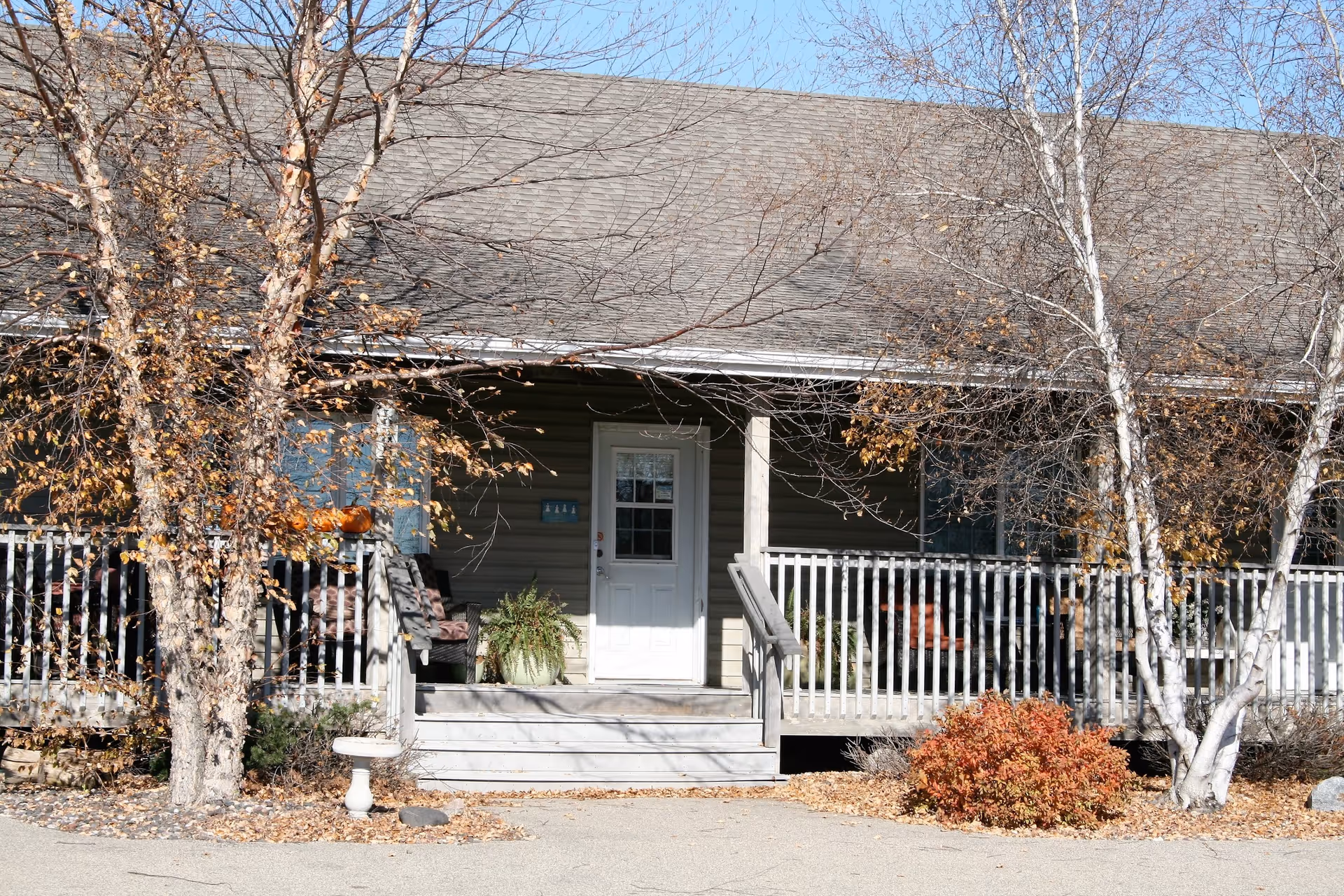 Front porch entrance of a single-story lodge with a white door, wooden railing, steps, and leafless trees and shrubs in front.