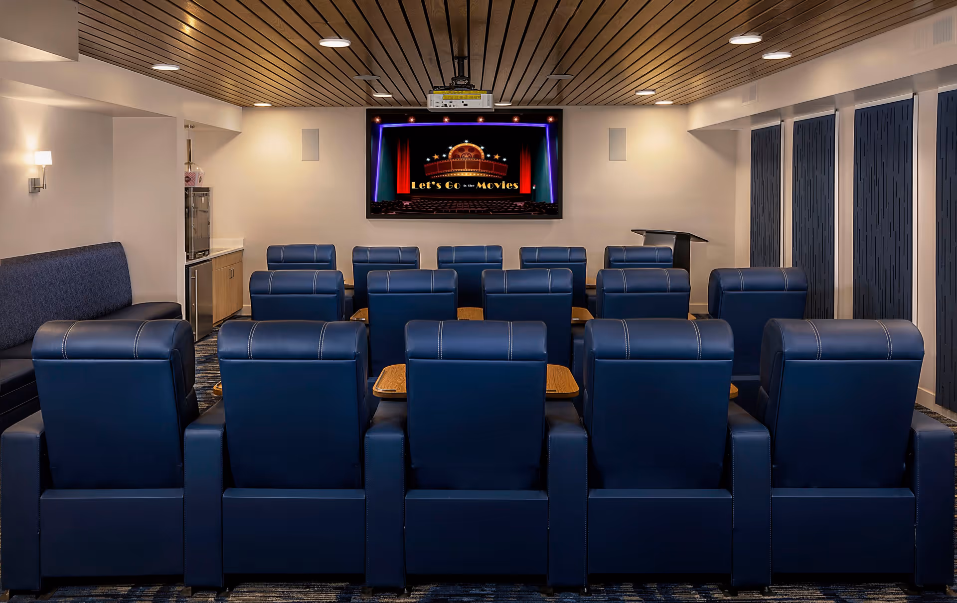 A small movie theater room with three rows of blue leather recliner chairs facing a large screen displaying the message 'Let's Go to the Movies'. The room has a wooden slatted ceiling, wall sconces for lighting, and a podium on the right side. There is also a popcorn machine and a small counter area on the left side.