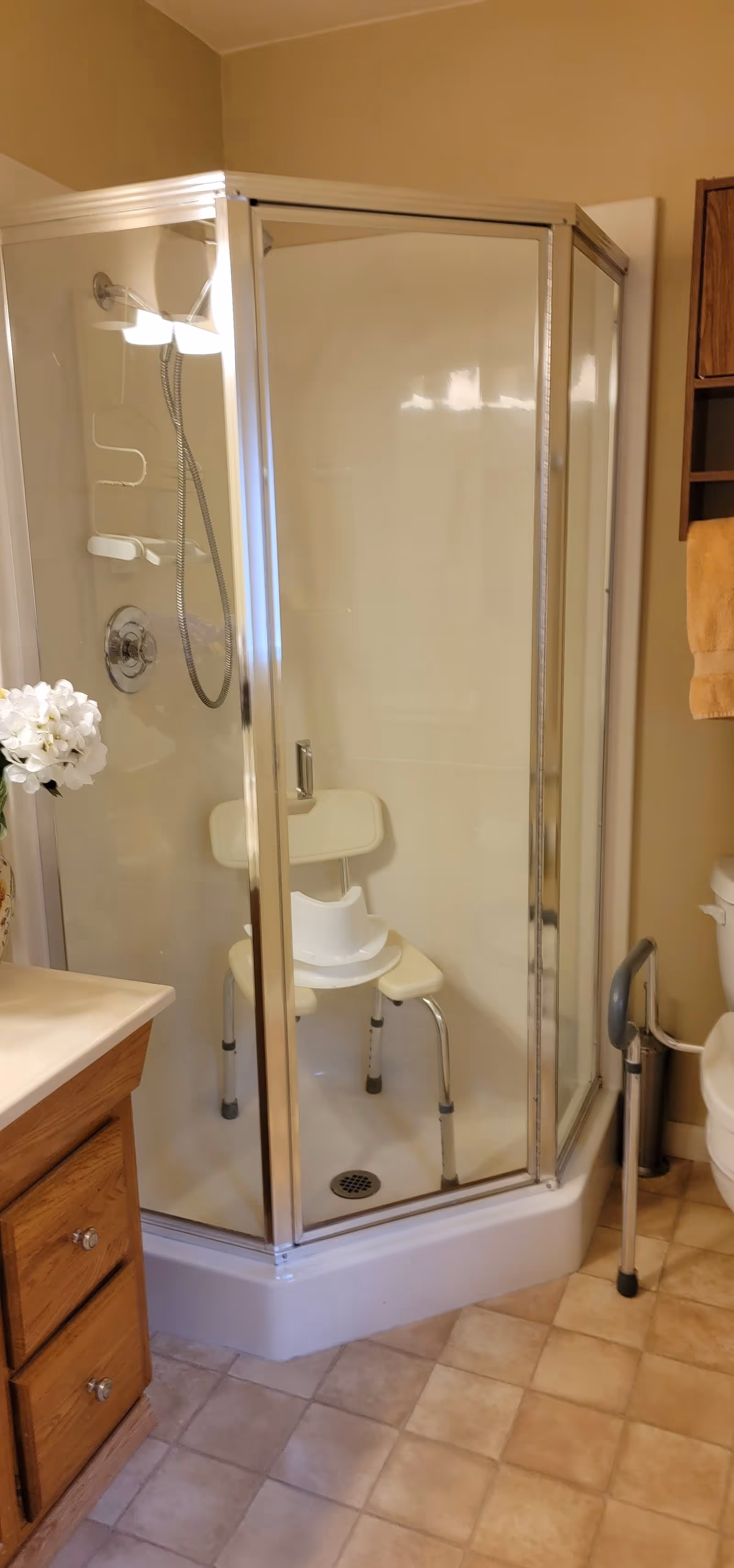 A bathroom with a corner glass shower enclosure containing a white shower chair. The bathroom has beige walls, a wooden vanity with a white countertop and a vase of white flowers, a toilet with a safety rail, and a towel hanging on a wooden cabinet.