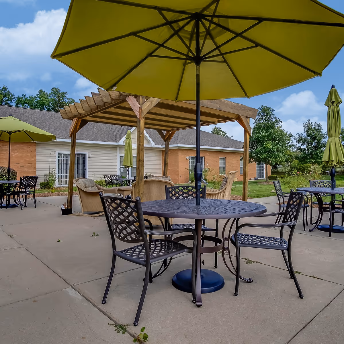 Outdoor patio area with metal tables and chairs, large green umbrellas, and a wooden pergola with cushioned seating. The background shows a single-story building with brick and siding exterior, surrounded by trees and greenery under a partly cloudy sky.