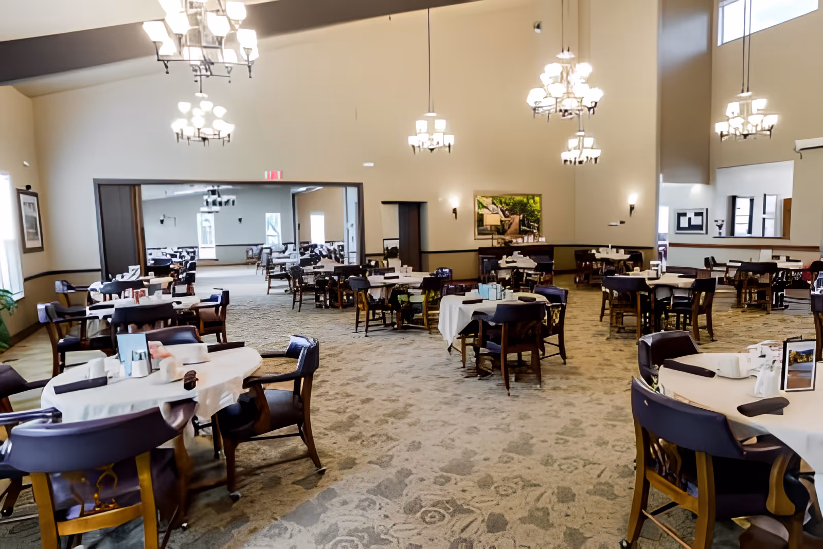 Spacious dining room with multiple round tables covered with white tablecloths and surrounded by wooden chairs with dark cushions. The room features high ceilings with several chandeliers providing warm lighting. There is a large open doorway leading to another dining area with similar setup. The walls are light-colored with framed artwork and windows allowing natural light.