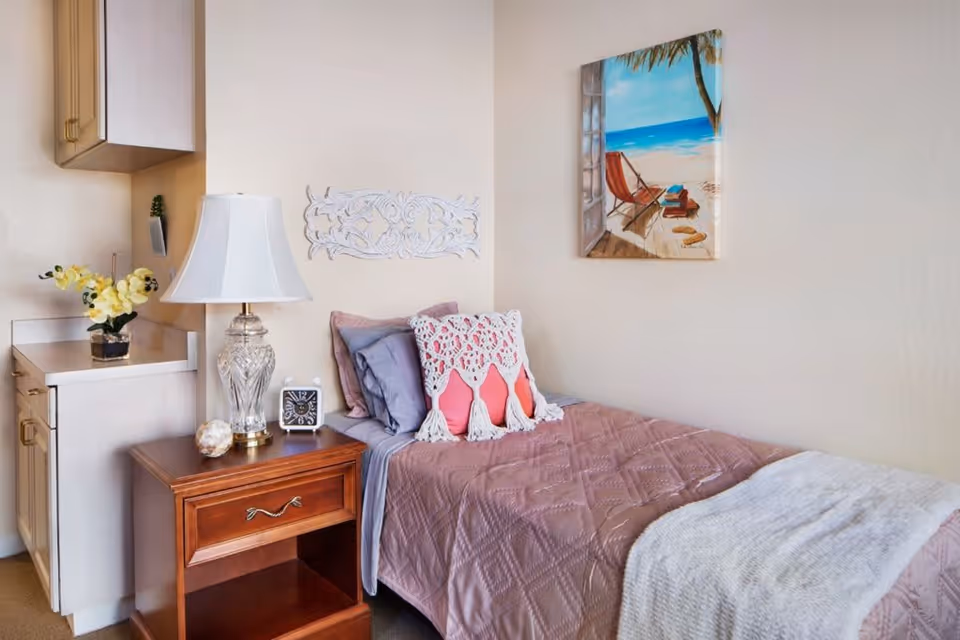A cozy bedroom featuring a single bed with mauve bedding, decorative pillows, and a light gray throw blanket. Next to the bed is a wooden nightstand with a glass lamp, a small clock, and a decorative item. On the wall above the bed is a painting of a beach scene with a chair, ocean, and palm tree. To the left, there is a small countertop with a vase of yellow flowers and a cabinet above it.