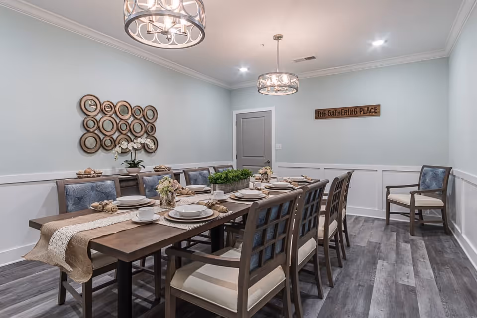 A dining room with a long wooden table set for a meal with plates, cups, and napkins. The room has light blue walls with white wainscoting, two modern circular chandeliers, and a decorative wall piece with multiple round mirrors. A wooden sign on the wall reads 'THE GATHERING PLACE'. There are cushioned chairs around the table and one chair against the wall.