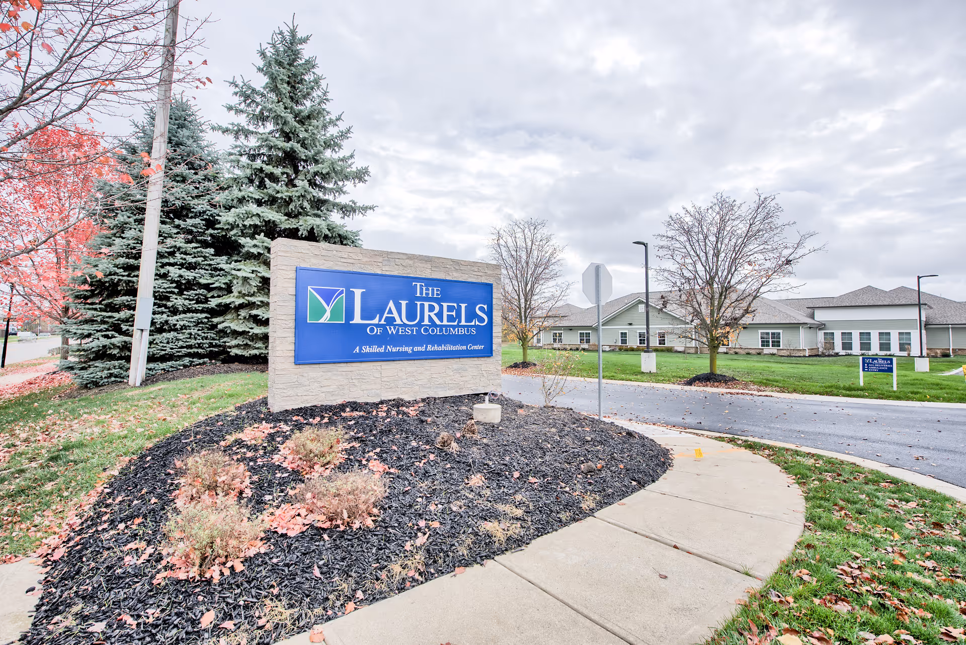 Outdoor view of the entrance area to The Laurels of West Columbus, featuring a large stone sign with the facility's name and description as a skilled nursing and rehabilitation center. The scene includes a curved sidewalk, landscaped mulch beds with small bushes, trees with autumn leaves, and a building in the background under a cloudy sky.
