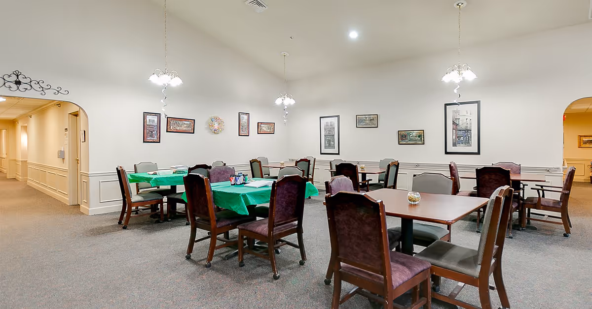 Communal dining room with multiple tables and wooden chairs, some tables covered with green tablecloths, framed artwork on the walls and hanging pendant lights.