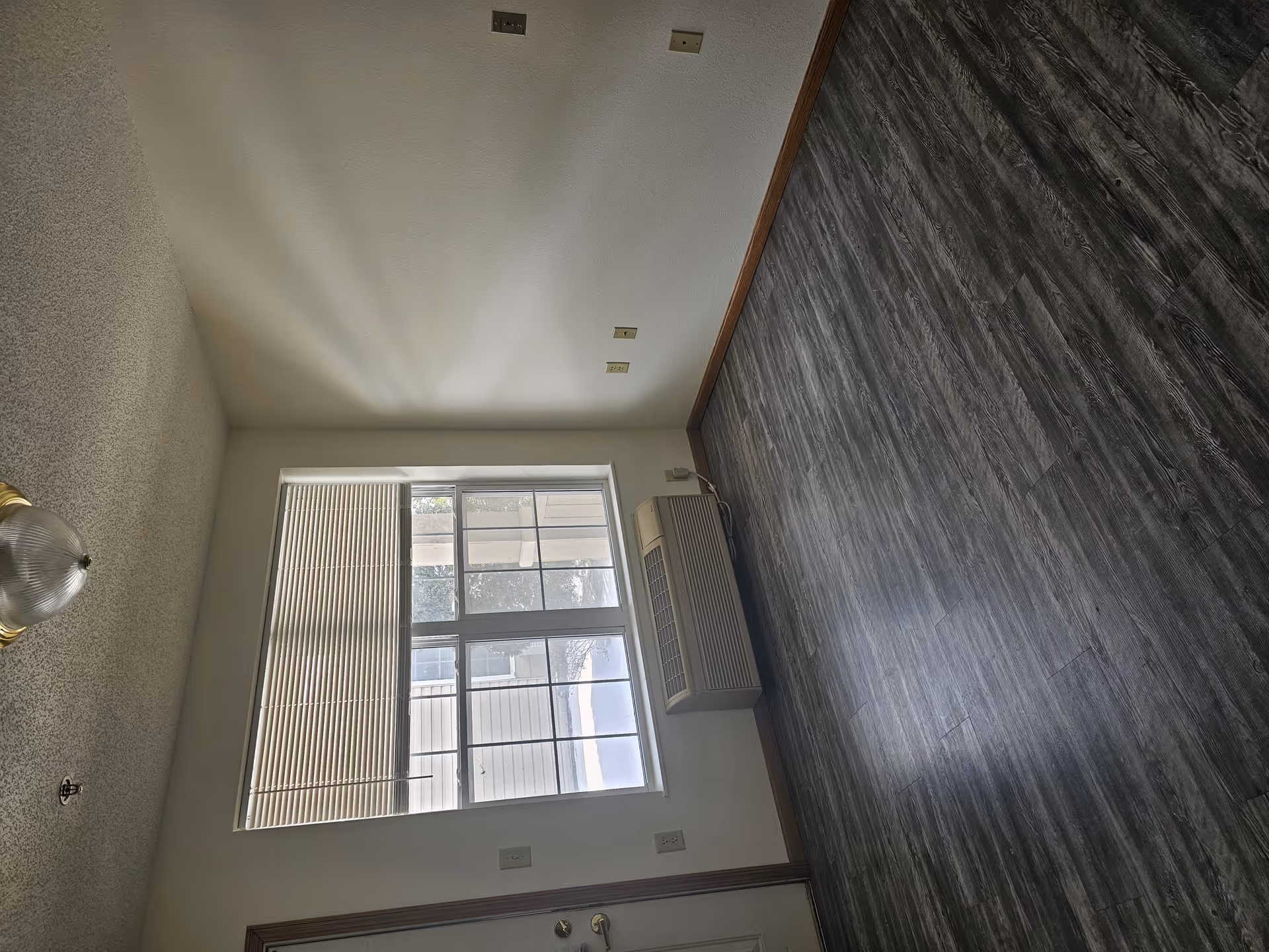 Empty room with gray wood-patterned flooring, white walls, a window with closed blinds, a wall-mounted air conditioning unit below the window, and a ceiling light fixture.
